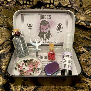 Könnte beinhalten: Eine silberne Blechdose mit schwarzem Deckel, die ein Hades-thematisches Hexenset enthält. Das Set beinhaltet eine kleine Flasche mit schwarzem Sand, eine kleine Flasche mit roten Kristallen, eine kleine Flasche mit schwarzer Flüssigkeit, einen silbernen Anhänger mit einem Symbol, einen violetten Kristall und einen kleinen Behälter mit rosa und weißen Kristallen.