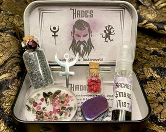 May include: A silver tin box with a black lid containing a Hades themed witchcraft kit. The kit includes a small bottle of black sand, a small bottle of red crystals, a small bottle of black liquid, a silver charm with a symbol, a purple crystal, and a small container of pink and white crystals.