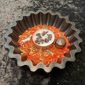May include: A decorative metal bowl with a scalloped edge, filled with orange rice, herbs, and a small candle. A silver wolf charm and a brown stone are also inside. The candle is decorated with small, dark stones.