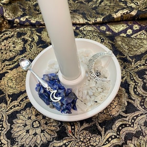 May include: A white ceramic candle holder with a central pillar and three compartments. One holds blue stones, another white stones, and the third a silver crescent moon charm. A small silver spoon rests on the edge.