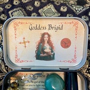 May include: An open, rectangular tin featuring a printed image of Goddess Brigid with red hair and a green dress. The tin contains a green stone, a small bottle, and other items. The text "Goddess Brigid" is visible on the lid.