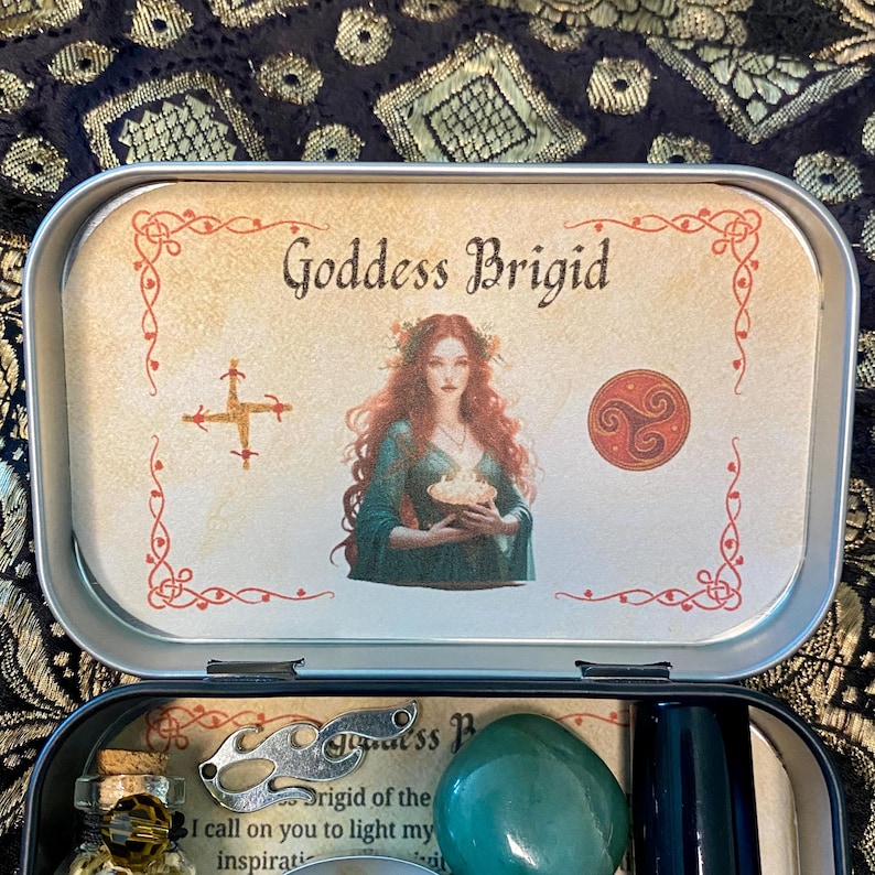 May include: An open, rectangular tin featuring a printed image of Goddess Brigid with red hair and a green dress. The tin contains a green stone, a small bottle, and other items. The text "Goddess Brigid" is visible on the lid.