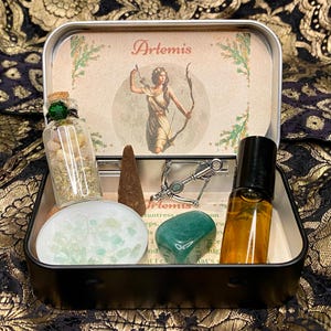 May include: A silver tin box with a green and brown image of Artemis, the Greek goddess of the hunt, holding a bow and arrow. The box contains a small bottle of herbs, a green crystal, a silver charm, a brown incense cone, and a small bottle of essential oil.