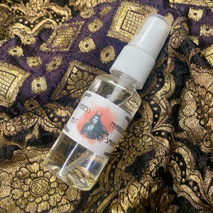 May include: A clear glass bottle with a white spray top and a label that reads "Goddess Mist" and "Magickal Spray". The bottle is sitting on a gold and black fabric.