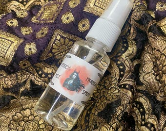 May include: A clear glass bottle with a white spray top and a label that reads "Goddess Mist" and "Magickal Spray". The bottle is sitting on a gold and black fabric.