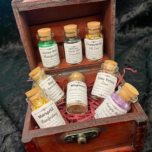 May include: A wooden box filled with small glass jars containing various herbs and crystals. Each jar has a label with a name and description, such as "Green Rice Manifesting", "Witches Black Salt Protection", "Chamomile Self Care", "Sage Banishing", "Mugwort Moon Magic", "Rose Petals Love", and "Purple Salt Spiritual Awakening".