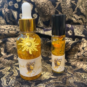 May include: Two bottles of golden yellow oil with a sun charm and a bust of Apollo on the label. The oil is in a clear glass bottle with a gold cap and a dropper. The smaller bottle is a roll-on.