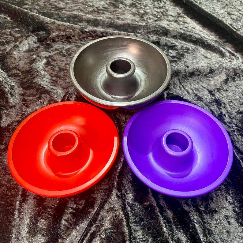 May include: Three colorful candle holders in red, purple, and gray. Each holder has a circular base with a raised center for a candle. The holders are arranged on a dark, textured surface.
