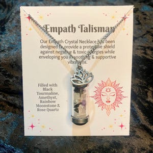 May include: A silver necklace featuring a clear vial pendant filled with crystals. The pendant has a lotus flower design at the top. The card behind the necklace reads "Empath Talisman" and lists the crystals: Black Tourmaline, Amethyst, Rainbow Moonstone & Rose Quartz.