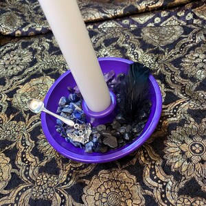 May include: A purple candle holder with a white candle, small silver spoon, and a black feather. The holder contains blue and black stones, and a silver bat charm. The background is a black and gold floral pattern.