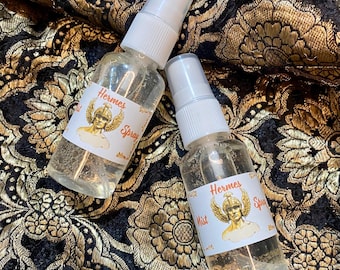 May include: Two clear spray bottles with white tops, labelled "Hermes Spray" and "Hermes Mist", featuring a gold illustration of a winged figure. The bottles are on a black and gold floral patterned fabric.