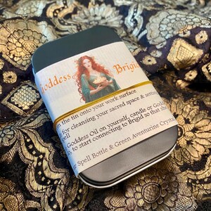 May include: A dark green rectangular tin with a label featuring the text "Goddess Brigid" and an illustration of a woman. The label is secured with a gold elastic band. The tin contains a spell bottle and green aventurine crystal.