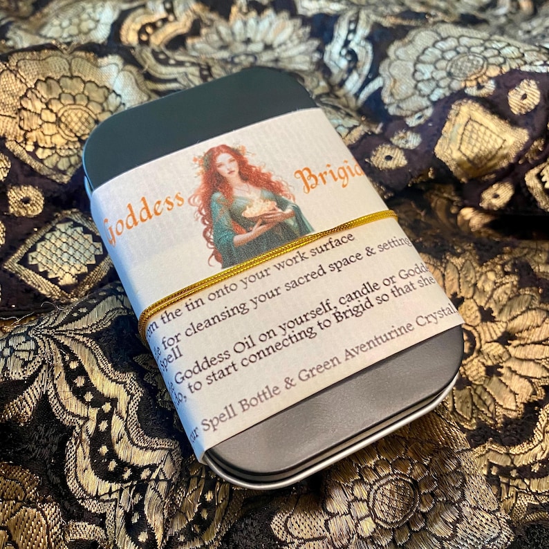 May include: A dark green rectangular tin with a label featuring the text "Goddess Brigid" and an illustration of a woman. The label is secured with a gold elastic band. The tin contains a spell bottle and green aventurine crystal.