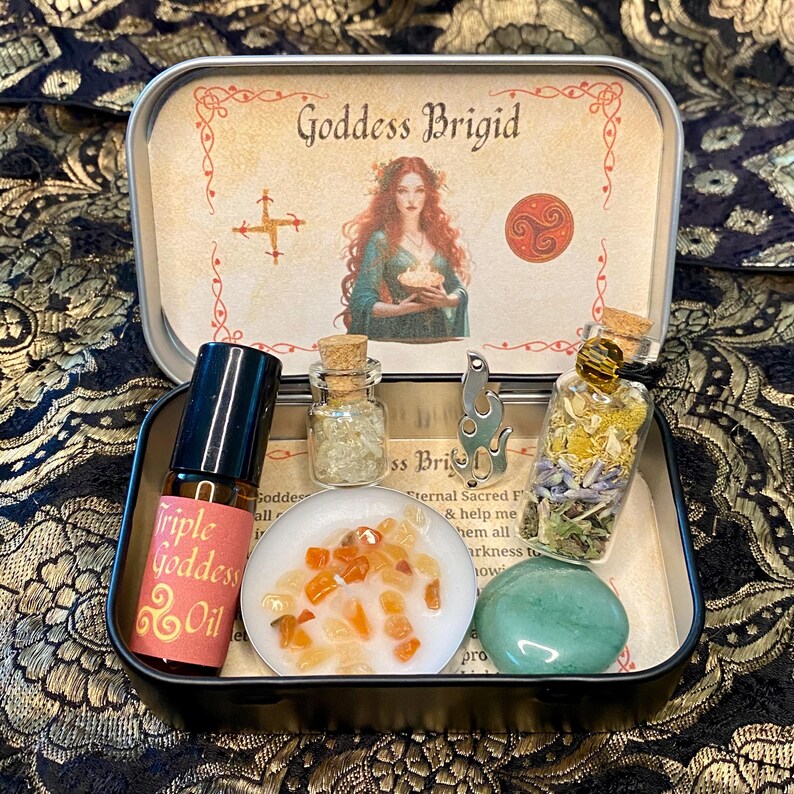 May include: A black metal tin with the words "Goddess Brigid" and an illustration of a woman. Inside are small bottles, a stone, a candle, and a bottle of "Triple Goddess Oil". The tin is on a patterned surface.