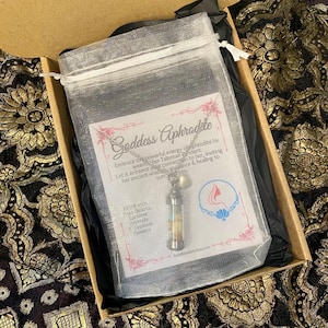 May include: A silver necklace with a pendant in a clear vial, filled with rose quartz, larimar crystals, and jasmine flowers, inside a mesh bag. The bag is in a box with the text "Goddess Aphrodite".