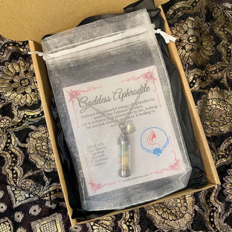 May include: A silver necklace with a pendant in a clear vial, filled with rose quartz, larimar crystals, and jasmine flowers, inside a mesh bag. The bag is in a box with the text "Goddess Aphrodite".