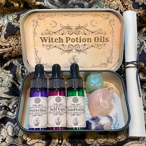 May include: A metal tin labeled "Witch Potion Oils" contains three glass dropper bottles with purple, pink, and green liquids. The bottles are labeled "Protection", "Self Care", and "Manifesting". Also included are crystals and a scroll.