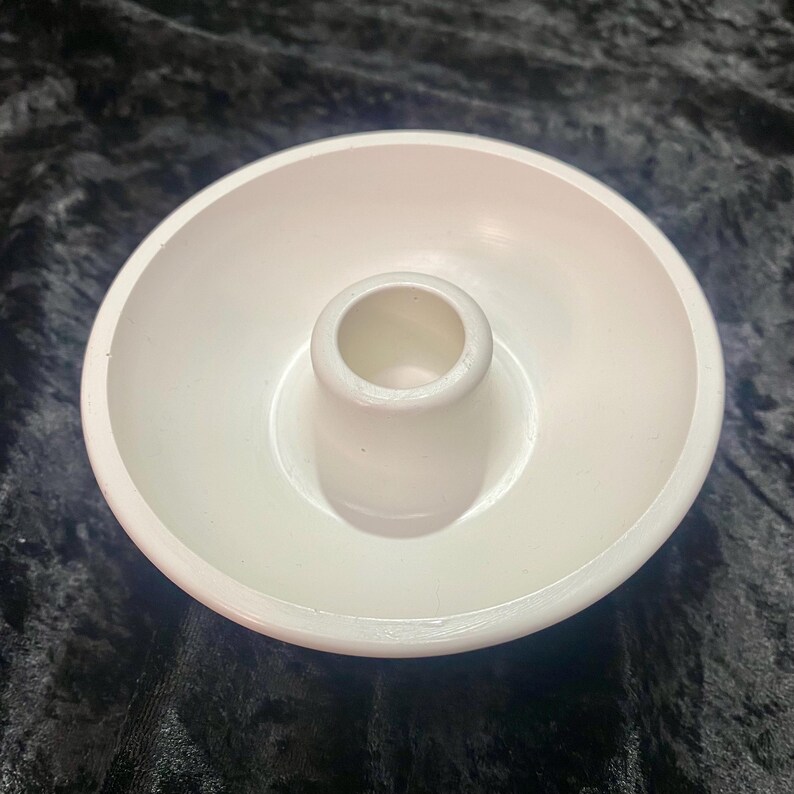 May include: A white ceramic candle holder with a circular base and a central, cylindrical cup for holding a candle. The holder has a simple, minimalist design.
