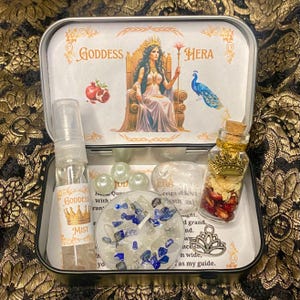 May include: A decorative metal tin featuring the words "Goddess Hera" and an illustration of a seated woman. The tin contains a spray bottle, a candle with blue stones, a small jar with a crown, and other decorative items.