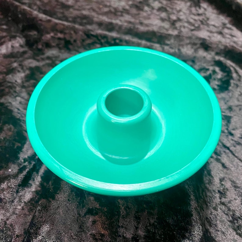 May include: A bright teal candle holder with a circular base and a central, cylindrical opening for a candle. The holder has a smooth, glossy finish and is set against a dark, textured background.