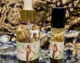 Puede incluir: Dos frascos de perfume Goddess de vidrio. El frasco más grande tiene una tapa dorada y un cuentagotas blanco. Un dije plateado de un carnero cuelga del cuello. La etiqueta presenta una ilustración de una mujer con arco y flecha. El frasco más pequeño tiene una tapa negra.