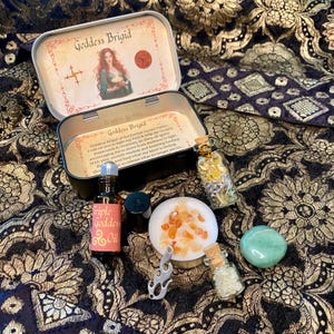 May include: An open Goddess Brigid tin with an illustration of a woman, surrounded by various spiritual items. These include small glass bottles, a small dish of orange crystals, a green stone, and a bottle of Triple Goddess Oil.