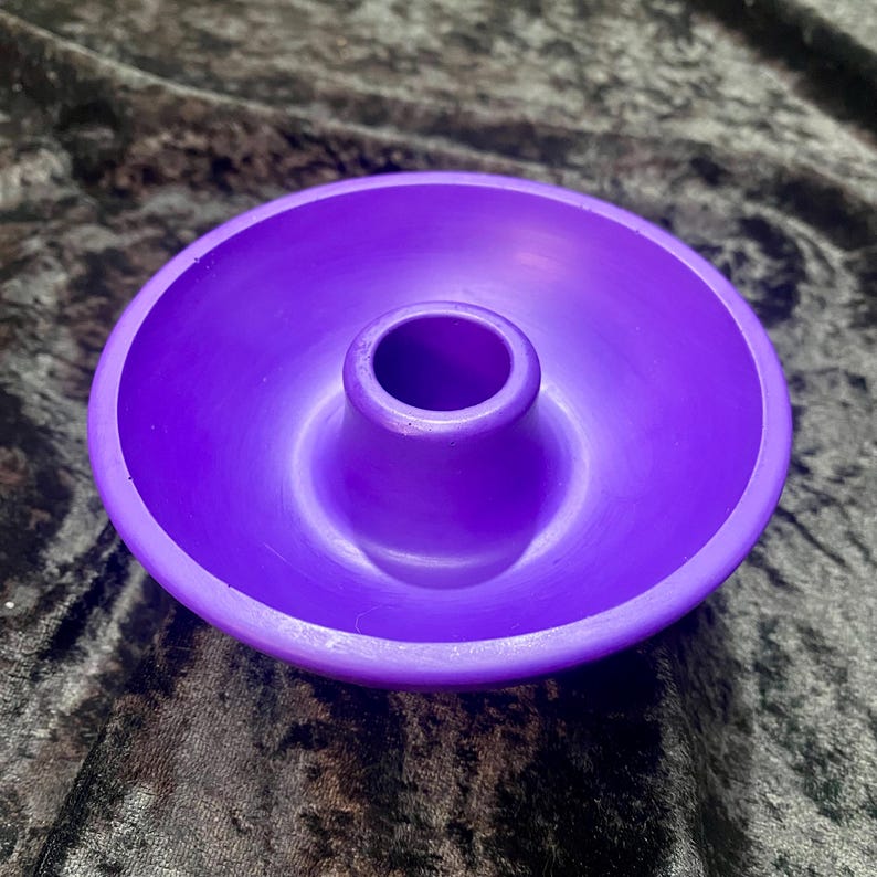 May include: A vibrant purple candle holder with a circular base and a central, raised cylinder. The holder has a smooth, matte finish and is designed to hold a single candle. The overall design is simple and modern.