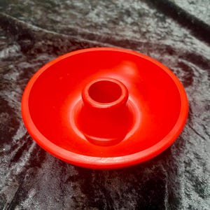 May include: A bright red candle holder with a circular base and a central, cylindrical cup. The holder is made of a smooth, solid material and is designed to hold a single candle. The design is simple and modern.