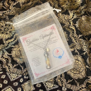 May include: A clear, drawstring bag containing a pendant necklace with a small glass vial filled with crystals and flowers. The bag also holds a card with the text "Goddess Aphrodite" and other text.