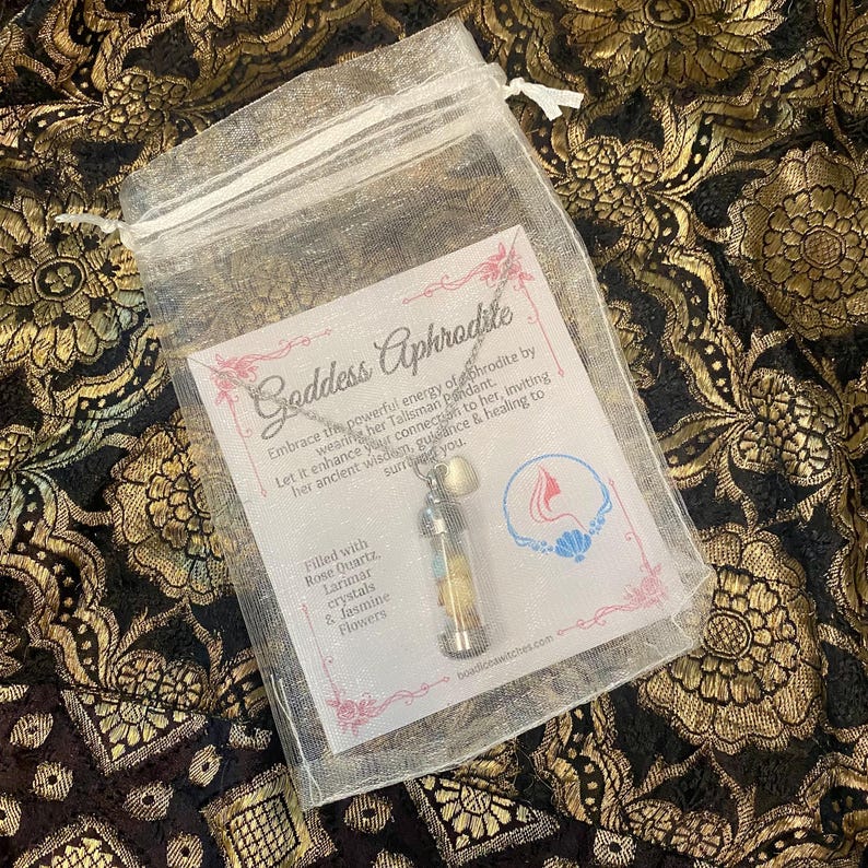 May include: A clear, drawstring bag containing a pendant necklace with a small glass vial filled with crystals and flowers. The bag also holds a card with the text "Goddess Aphrodite" and other text.