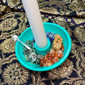 May include: A teal candle holder with a white candle. The holder contains blue, green, and orange gemstones. A small silver spoon and charm are also present. The holder sits on a patterned fabric.