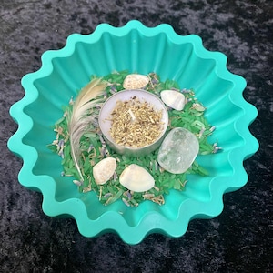May include: A teal, starburst-shaped dish filled with green rice, lavender, seashells, a feather, and a small candle. A clear quartz crystal sits next to the candle. The candle is surrounded by dried herbs.