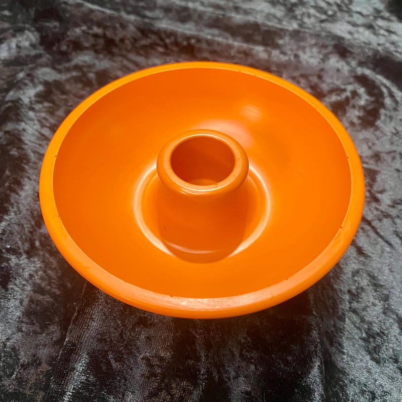 May include: Bright orange candle holder with a circular base and a central, cylindrical cup. The holder is made of a solid, glossy material. The design is simple and modern, suitable for various home decor styles.