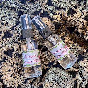 May include: Two clear spray bottles with black caps and labels reading "Dionysus Exilir Mist" in pink and green. They rest on a black and gold floral patterned fabric.