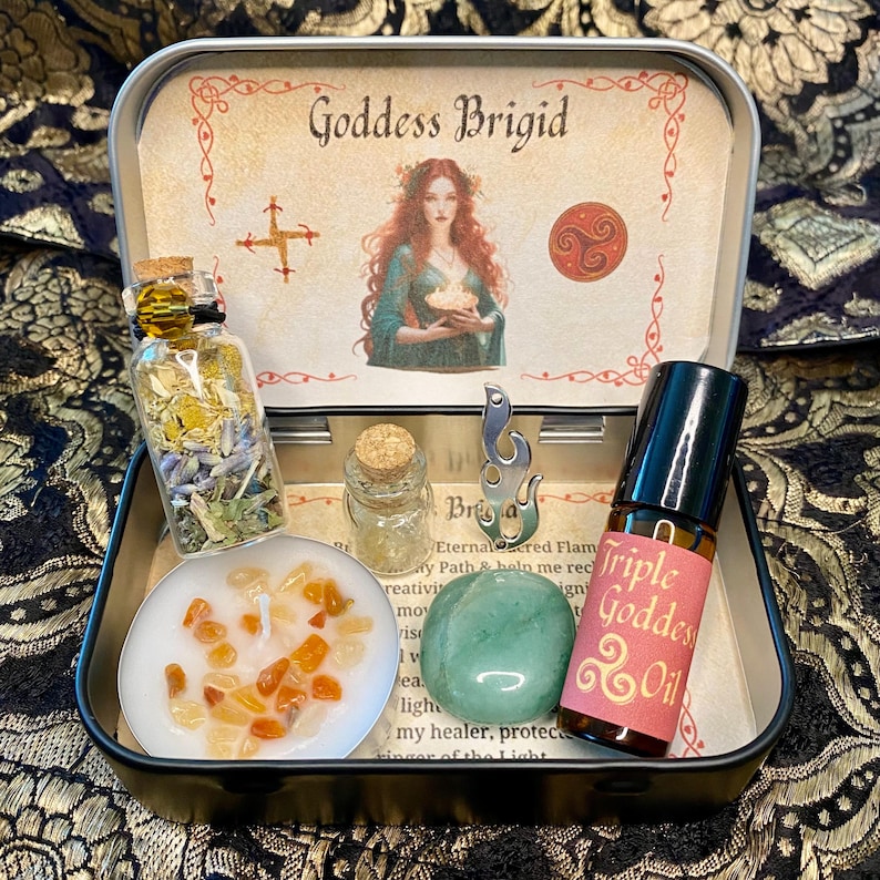 May include: An open black tin with the words "Goddess Brigid" and an illustration of a woman. Inside are small glass bottles, a candle with orange stones, a green stone, and a bottle of "Triple Goddess Oil".
