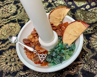 Pode incluir: Um queimador de incenso de cerâmica branca com um tubo central, rodeado por pedras laranja e verdes, fatias de maçã secas e uma pequena colher de prata com um pingente de nó celta. O queimador está sobre um tecido estampado.