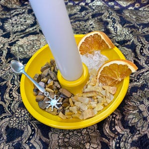 May include: A bright yellow candle holder with a white candle, dried orange slices, crystals, and small stones. A silver spoon with a sun-shaped charm is placed on the side. The items are on patterned fabric.