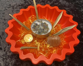 May include: A bright red, scalloped bowl containing orange rice, dried herbs, and a small candle with lavender. A gold coin and a brown stone are also present. The arrangement is set against a dark background.