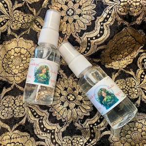 May include: Two clear spray bottles with white caps, labelled "Goddess Mist Gaia Spray", sit on black and gold patterned fabric. The labels feature an illustration of a woman with flowers in her hair.