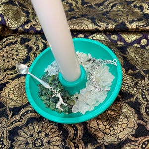 May include: A teal candle holder with a white candle, small silver spoon, and a crescent moon charm. The holder contains green and white stones. The background is a dark fabric with a gold floral pattern.