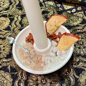 May include: A white candle holder with a white candle, dried apple slices, and various crystals. A small silver spoon and a charm with two connected hearts are also present. The arrangement is on a patterned surface.
