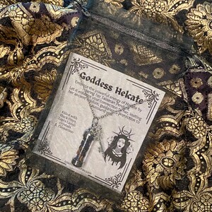 May include: A necklace with a pendant containing black onyx, lapis lazuli crystals, and cinnamon. The pendant hangs from a silver chain. The necklace is displayed on a patterned fabric with the text "Goddess Hekate".