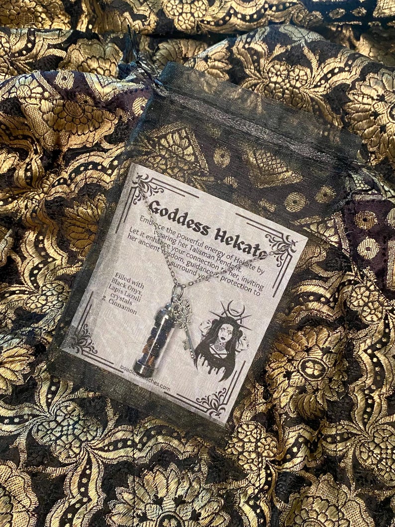 May include: A necklace with a pendant containing black onyx, lapis lazuli crystals, and cinnamon. The pendant hangs from a silver chain. The necklace is displayed on a patterned fabric with the text "Goddess Hekate".