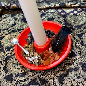 May include: A red candle holder with a white candle, black feather, and various stones. A silver spoon with a wolf charm rests on the edge. The holder is on a patterned fabric.