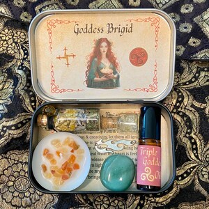 May include: An open metal tin featuring the text "Goddess Brigid" and an illustration of a woman. Inside are small glass bottles, a candle with orange stones, a green stone, and a bottle of "Triple Goddess Oil".