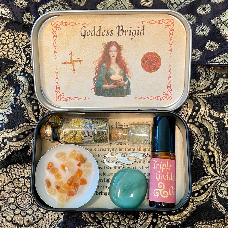 May include: An open metal tin featuring the text "Goddess Brigid" and an illustration of a woman. Inside are small glass bottles, a candle with orange stones, a green stone, and a bottle of "Triple Goddess Oil".
