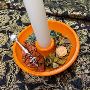May include: An orange candle holder with a white candle. The holder contains various coloured stones and a silver spoon with a medical symbol charm. A coin is also present.