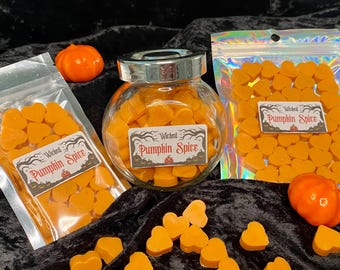 May include: Three glass jars filled with orange heart-shaped wax melts. The jars have labels that read "Wicked Pumpkin Spice".