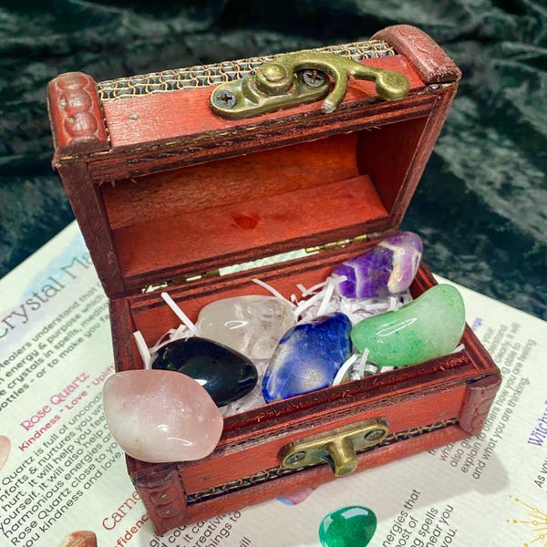 May include: A small, open wooden treasure chest with a brass clasp, filled with various colourful gemstones. The gemstones include rose quartz, amethyst, and green aventurine. The chest sits on a surface with printed text.