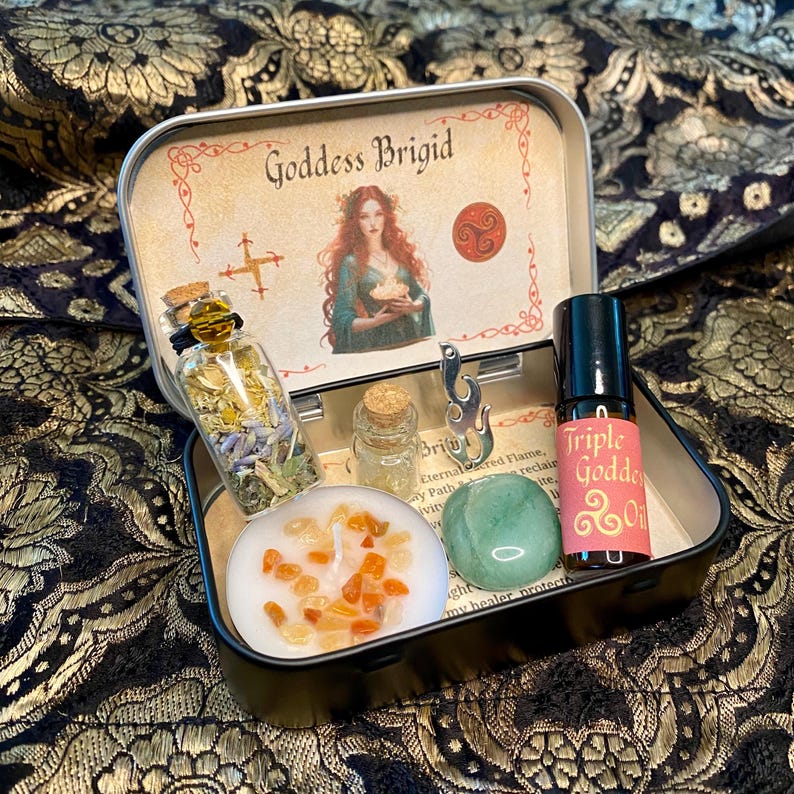 May include: A black metal Goddess Brigid kit with various items. Includes a small glass bottle with herbs, a candle with orange crystals, a green stone, a small bottle, a silver charm, and a bottle of Triple Goddess Oil.